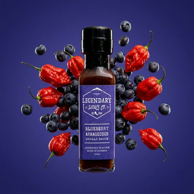 Bottle of Legendary Sauces Co. Blueberry Armageddon Chilli Sauce surrounded by blueberries and red chilli peppers on a purple background.